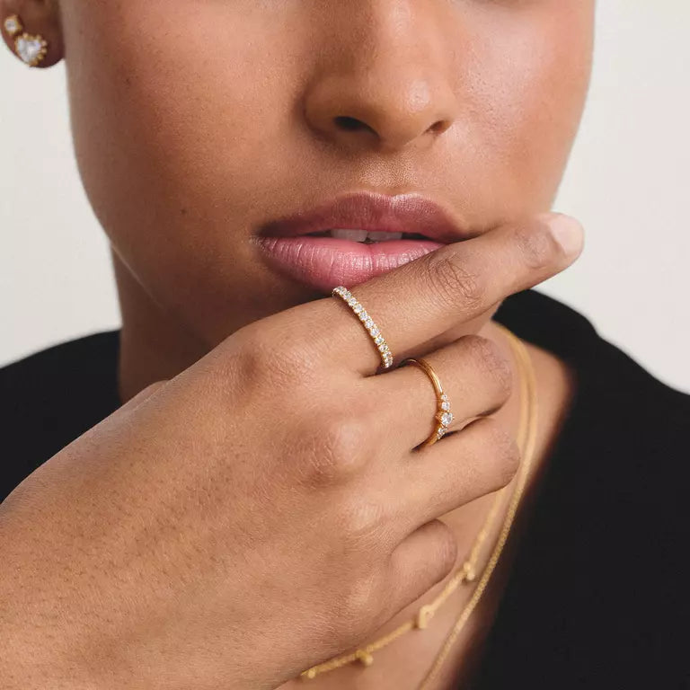Close-up of a person wearing two gold rings, one with a row of small clear stones and another with a single clear stone, along with a delicate gold necklace.