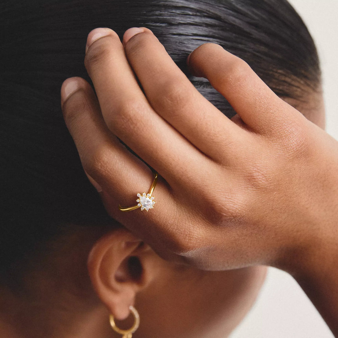 A close-up lifestyle shot of a person's hand resting on their head, wearing a delicate gold ring with a small clear gemstone in a flower-like setting. The person also wears a small gold hoop earring.