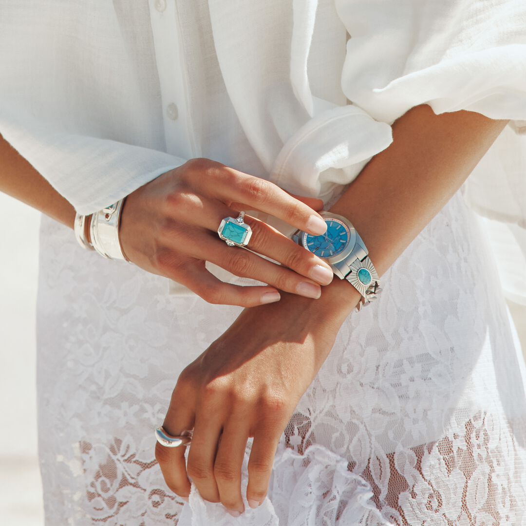 Close-up of a person's hands wearing a watch and rings with a white lace garment in the background