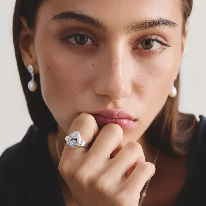 A close-up portrait of a woman wearing a silver ring with a marquise-cut clear gemstone on her finger, along with pearl drop earrings, set against a soft light background.