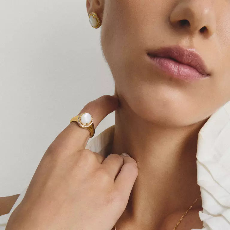 A close-up lifestyle shot of a model wearing a gold ring with a white pearl on her finger, along with a matching pearl stud earring and a white ruffled top.