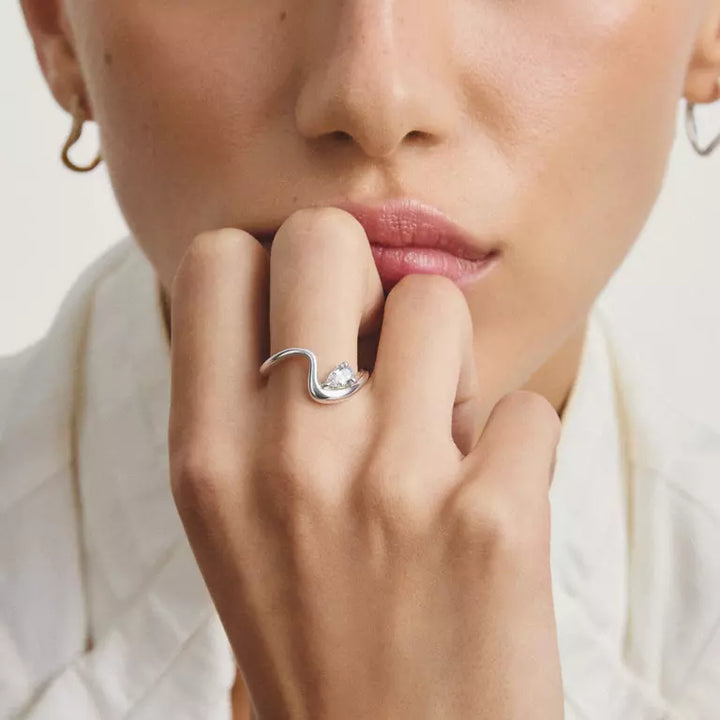 A close-up lifestyle shot of a model wearing a silver wavy ring with a clear central gemstone on her finger, paired with small hoop earrings and a white collared shirt.