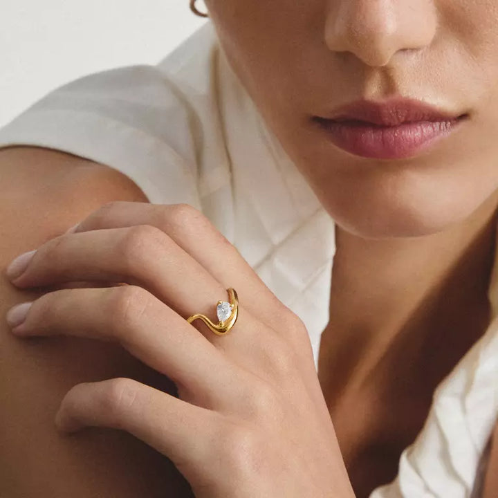 A close-up lifestyle shot of a model wearing a gold ring with a teardrop-shaped clear gemstone on her finger, set against a soft cream-colored top and a neutral background.