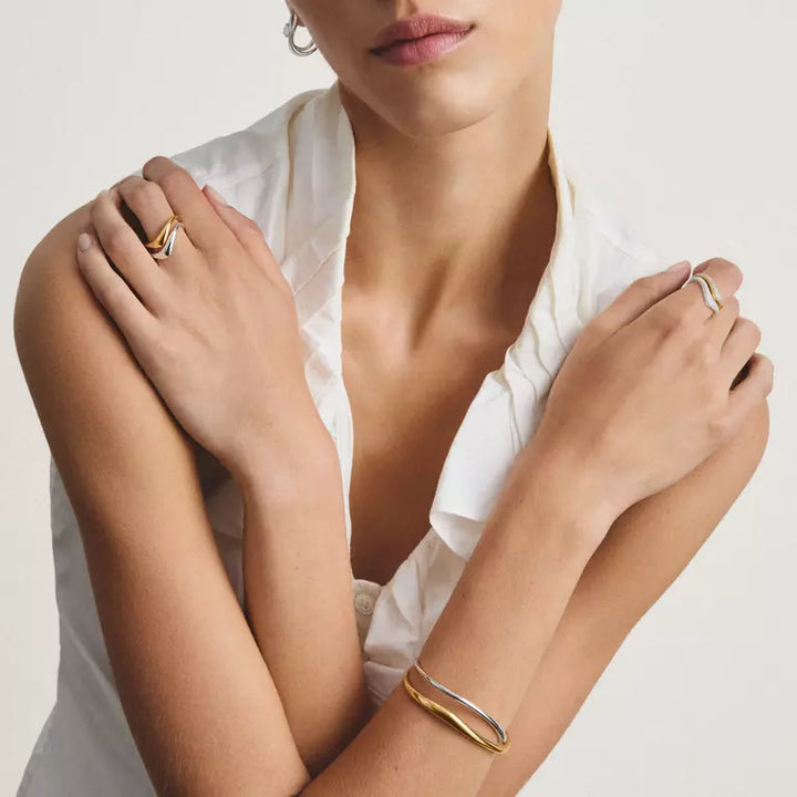 A close-up lifestyle shot of a model wearing minimalist gold and silver rings on her fingers and a matching two-tone bangle bracelet on her wrist, paired with a white blouse against a light beige background.