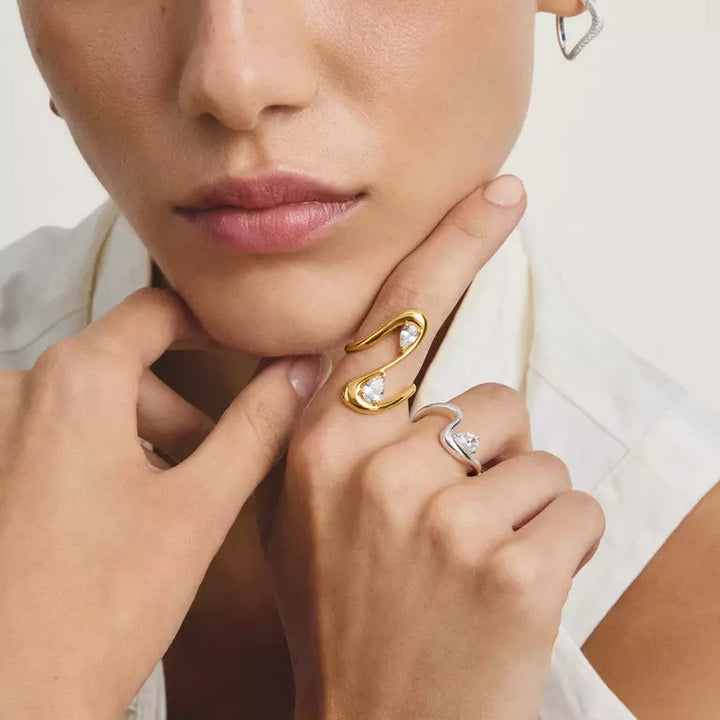 A close-up lifestyle shot of a model wearing two elegant rings on her fingers. The primary product is a gold ring featuring a curved, snake-like form with two small white pearl accents and a clear central gemstone.