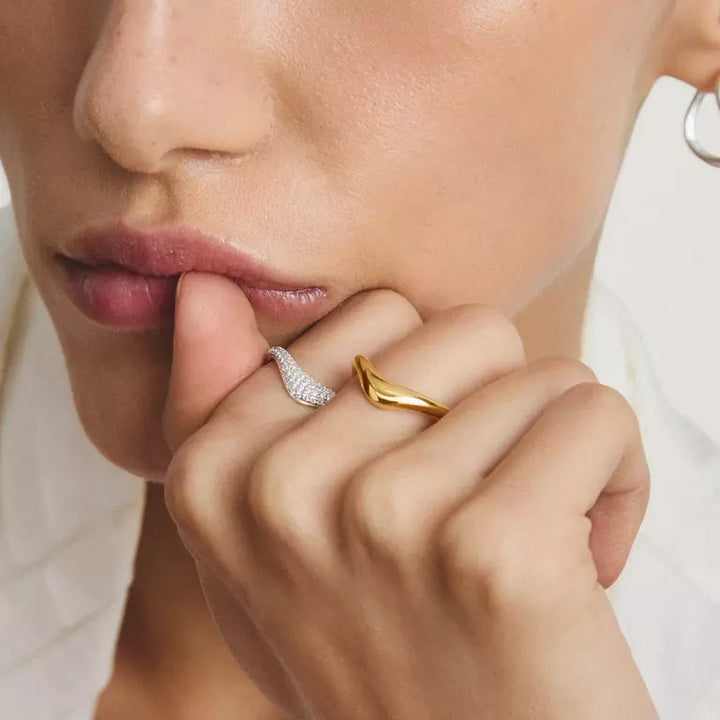Close-up of a person's hand and lower face wearing two slim wave-shaped rings, one in yellow gold and one in silver with small clear stones, against a soft light background.