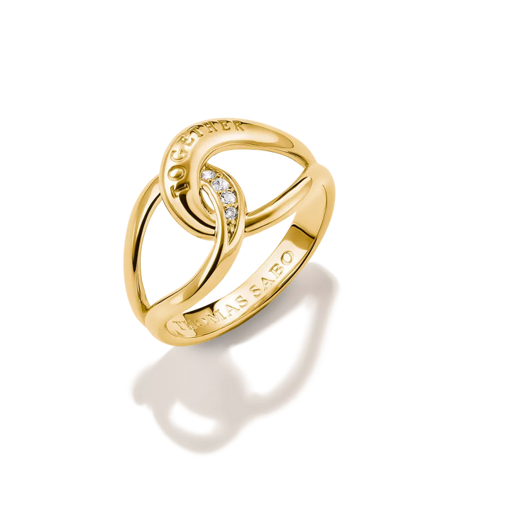 A gold interlocking rings ring with a small clear stone accent and 'TOGETHER' engraved on the top, and 'THMAS SABO' on the band, shown on a white background with a soft shadow.