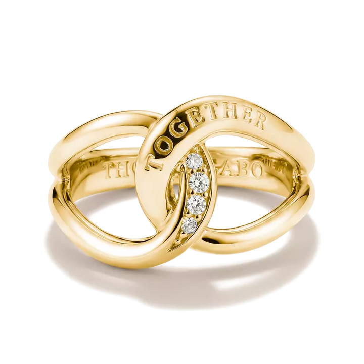 A gold interlocking rings ring with a small clear stone accent and 'TOGETHER' engraved on the top, and 'THMAS SABO' on the band, shown on a white background with a soft shadow.