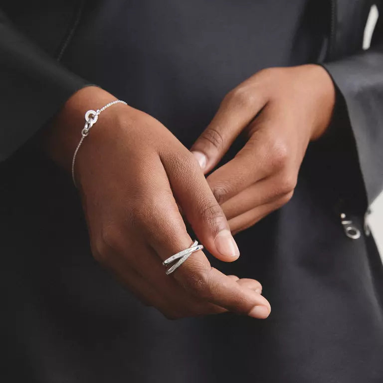 A close-up of a person's hands wearing a silver ring and a delicate silver bracelet, dressed in a black jacket against a dark background.