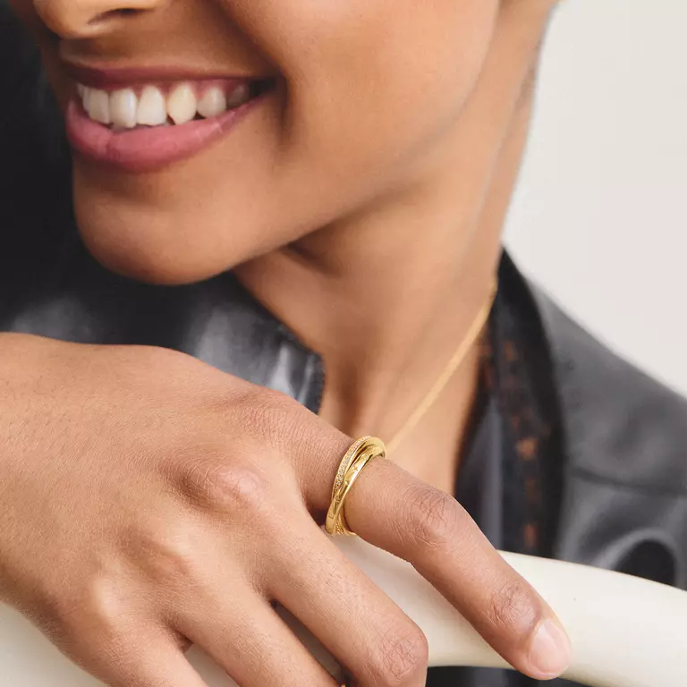 A close-up lifestyle shot of a model wearing a double-band gold ring on her finger, with a gold chain necklace also visible. She is wearing a black leather jacket against a light gray background.