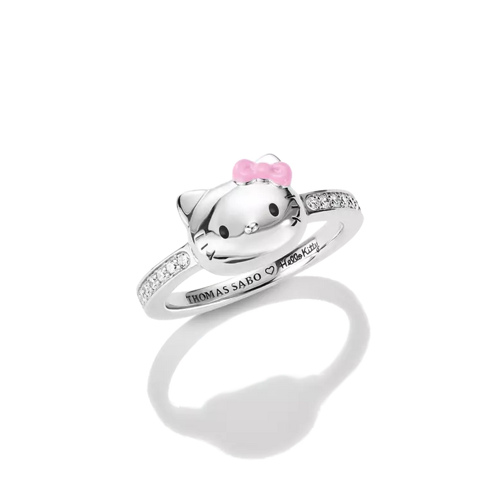 A silver Hello Kitty ring by Thomas Sabo, featuring a polished silver cat head with a pink bow, set on a silver band adorned with small clear stones, shown on a white background with a soft reflection.