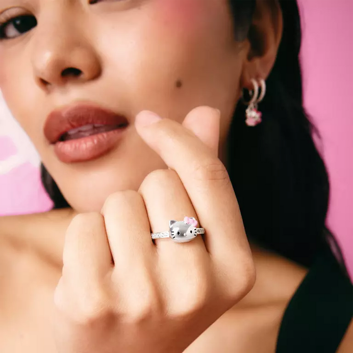 A close-up lifestyle shot of a model wearing a silver cat-face ring on her finger, with a matching pink hoop earring visible on her ear. The background is a solid vibrant pink.