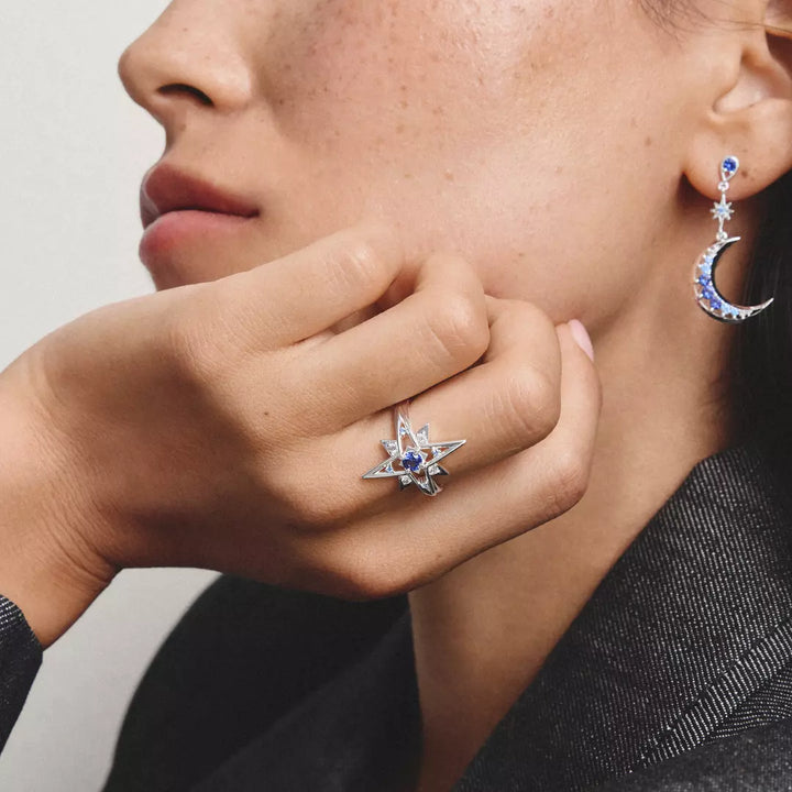 A close-up lifestyle shot of a model wearing a silver star ring with a blue gemstone and clear accent stones, alongside a matching crescent moon earring.