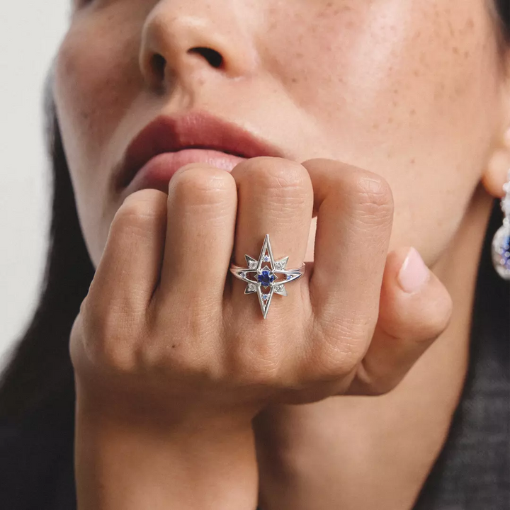 A close-up lifestyle shot of a model's hand wearing a silver ring with a starburst design, a central deep blue gemstone, and small clear accent stones.