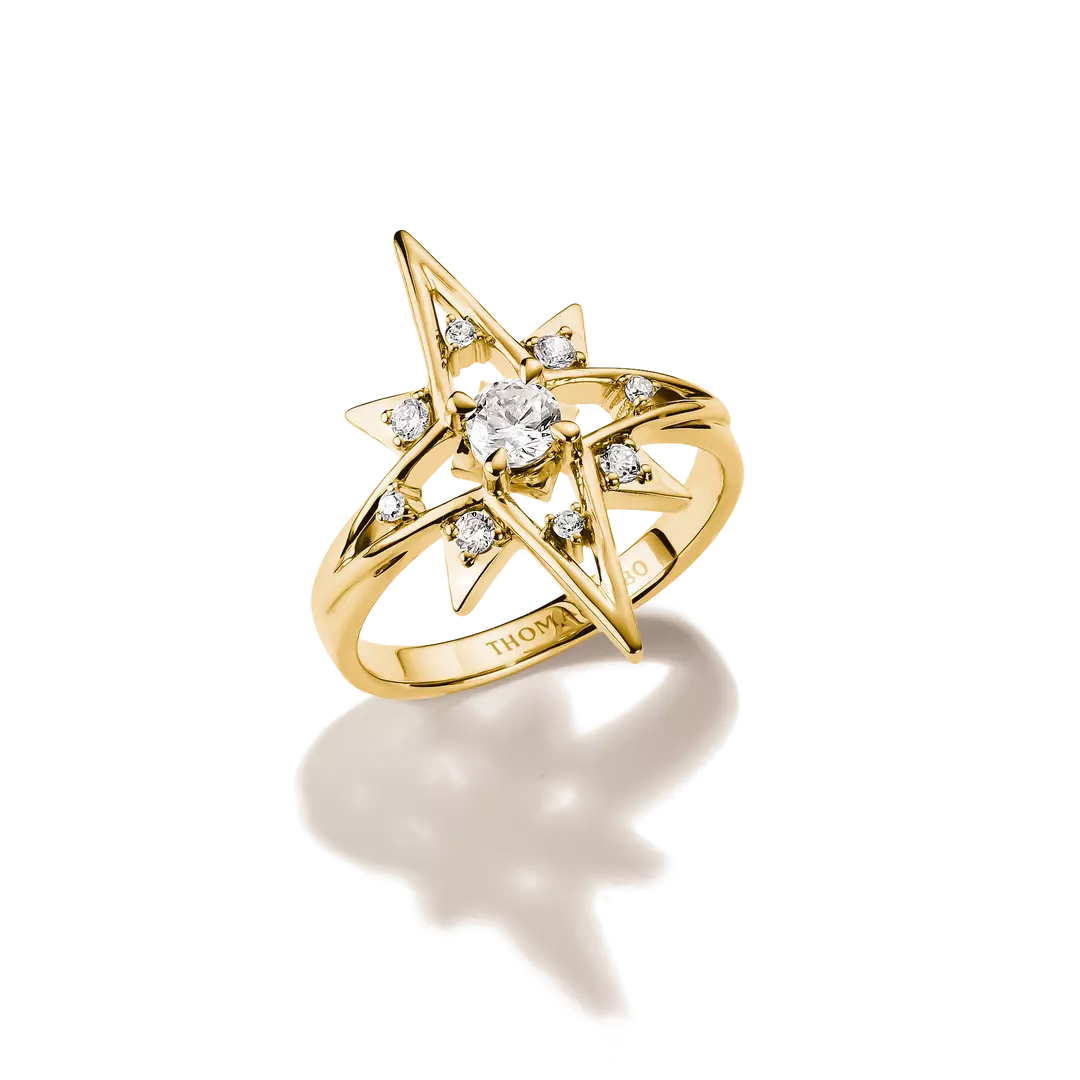 A gold ring featuring a starburst design with a central round clear gemstone and smaller surrounding clear stones, photographed on a clean white background with a soft shadow below.