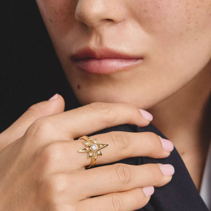 A close-up of a model's hand wearing a gold starburst ring with a central clear gemstone, set against a dark background.