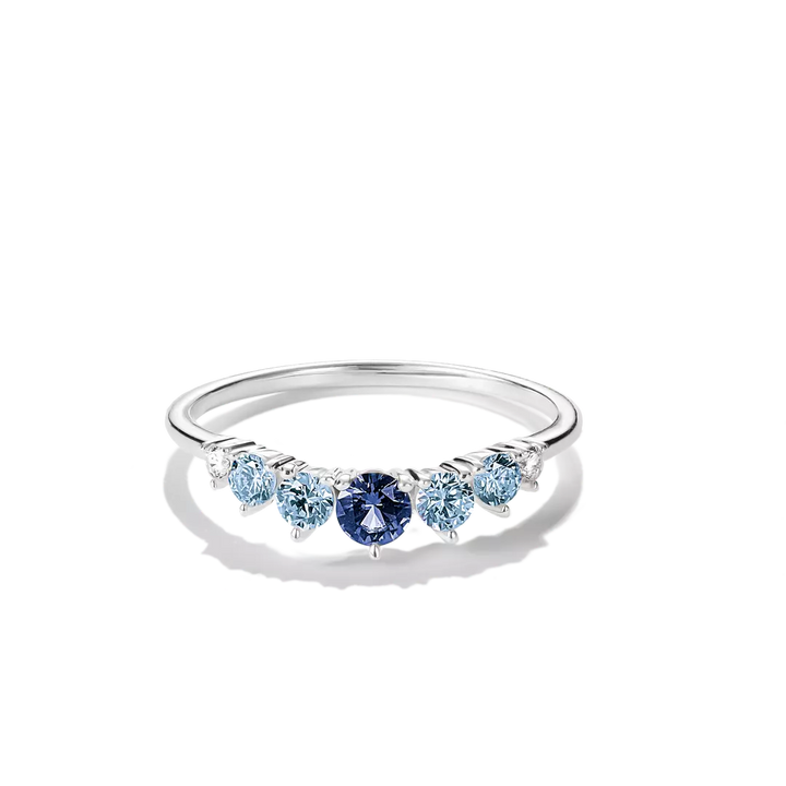 A silver band ring featuring a row of round-cut light blue and dark blue gemstones, displayed on a clean white background with a soft shadow.