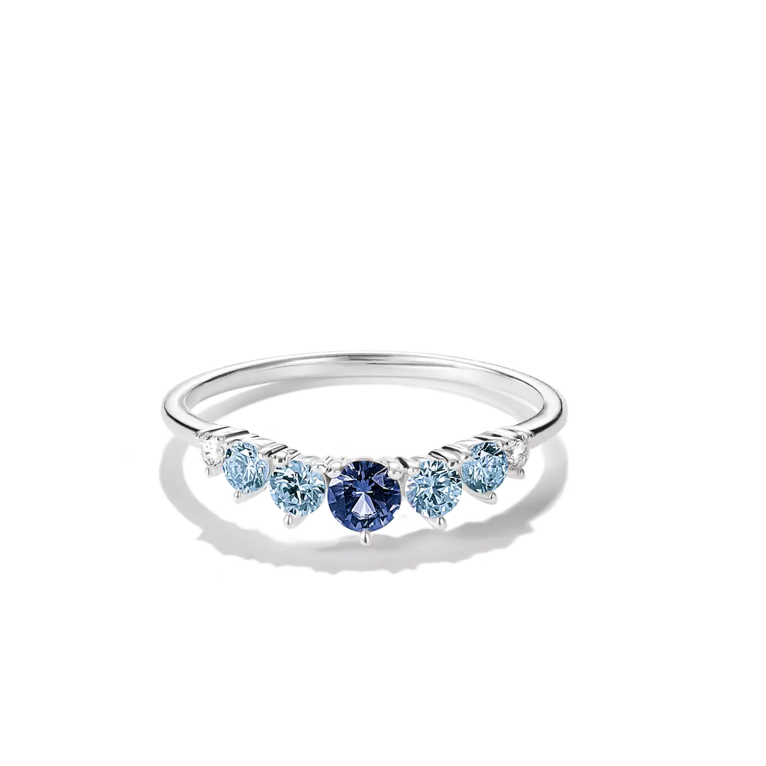 A silver band ring featuring a row of round-cut light blue and dark blue gemstones, displayed on a clean white background with a soft shadow.