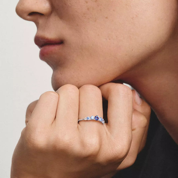 A close-up lifestyle shot of a person wearing a delicate silver band ring adorned with small clear stones and a central deep blue sapphire, resting their chin on their hand against a neutral light gray background.