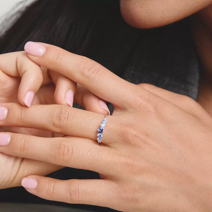 A close-up lifestyle shot of a delicate silver band ring with small blue gemstones, worn on a woman's ring finger.