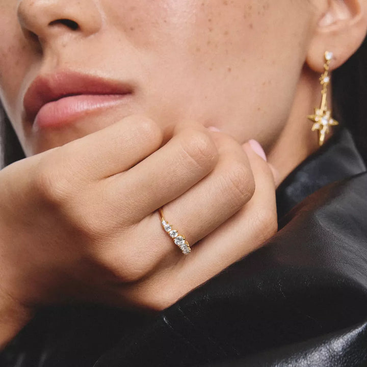 A close-up lifestyle shot of a model wearing a delicate gold ring with small clear stones on the ring finger, alongside a matching gold star-shaped earring.