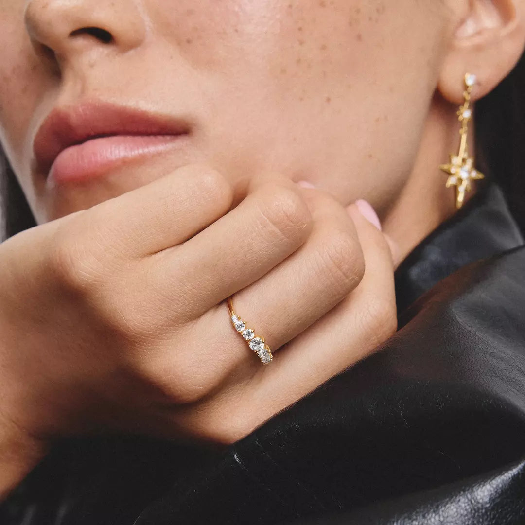 A close-up lifestyle shot of a model wearing a delicate gold ring with small clear stones on the ring finger, alongside a matching gold star-shaped earring.