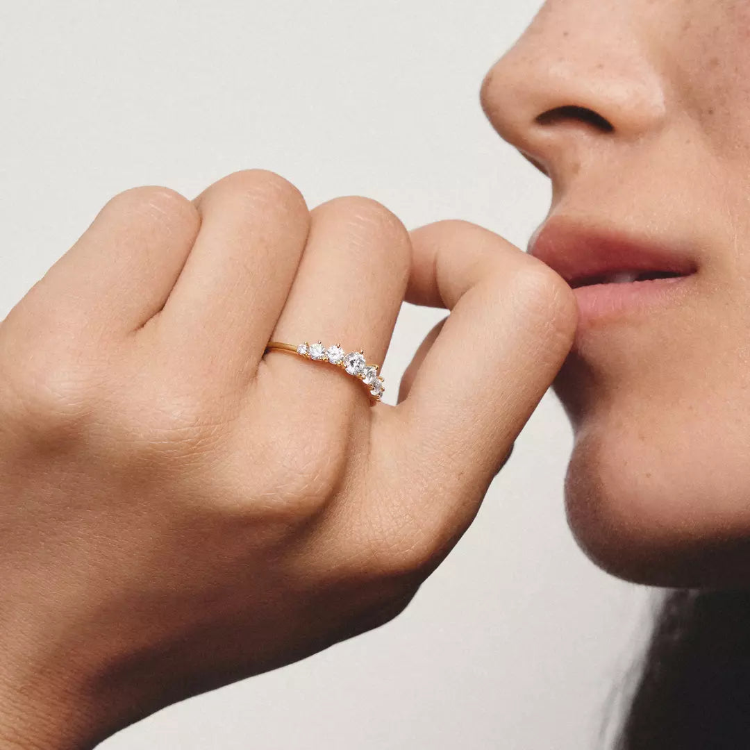 A close-up lifestyle shot of a person wearing a delicate gold ring with small clear gemstones on the ring finger, with their hand near their lips against a soft light gray background.