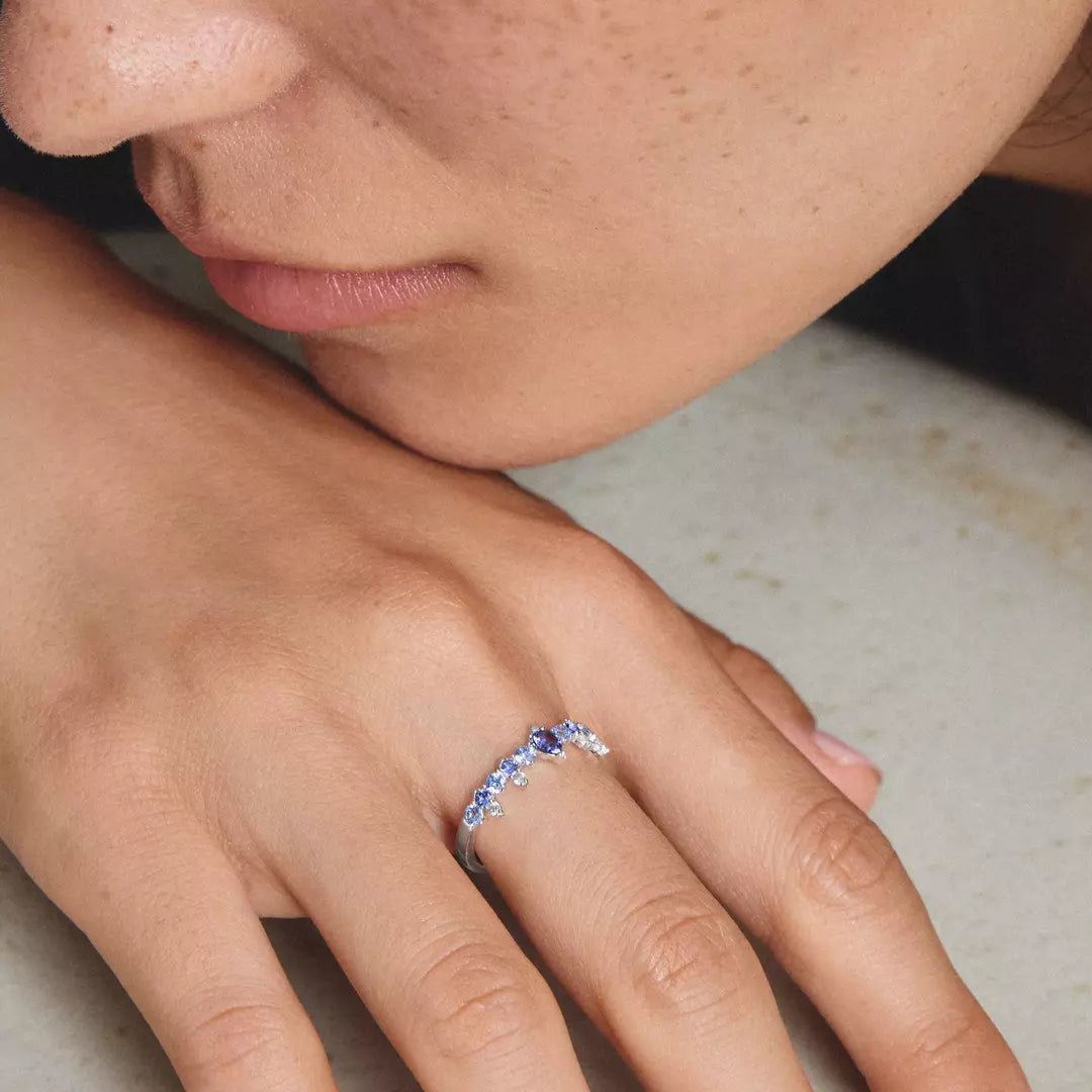 A close-up lifestyle shot of a person resting their head on their hand, showcasing a delicate silver ring with a central blue gemstone and small clear accent stones.