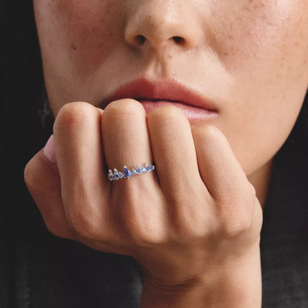 A close-up lifestyle shot of a person resting their head on their hand, showcasing a delicate silver ring with a central blue gemstone and small clear accent stones.
