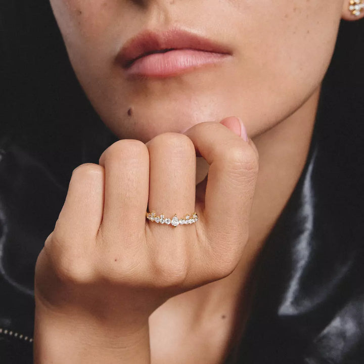 A close-up lifestyle shot of a model wearing a delicate gold ring with small clear stones on her ring finger. The model is wearing a black leather jacket, and the background is dark.