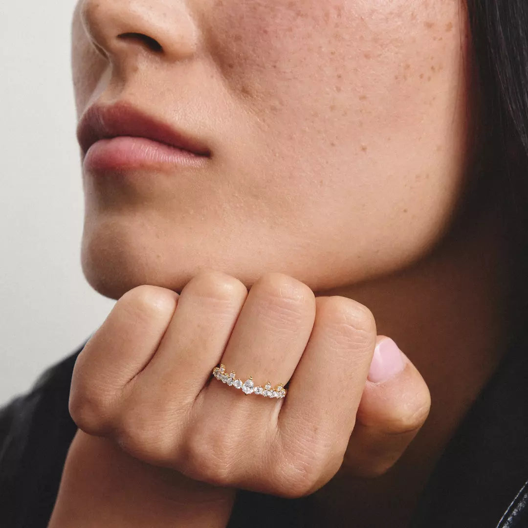 A close-up lifestyle shot of a model resting their chin on their hand, showcasing a delicate silver band ring adorned with small clear gemstones and tiny gold and white accent stones.